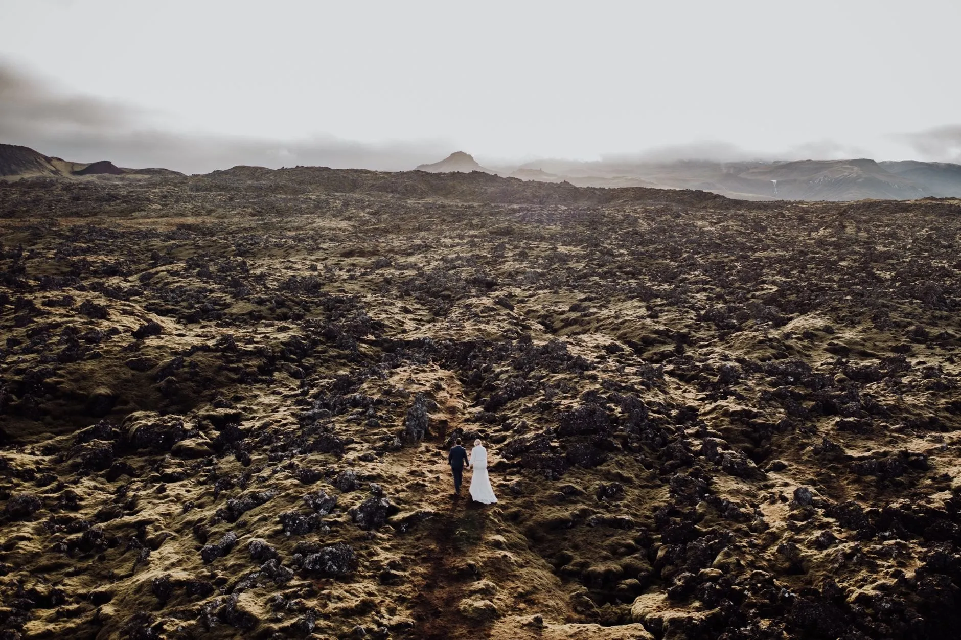 A couple walking hand in hand across a vast moss-covered lava field, tiny in the frame