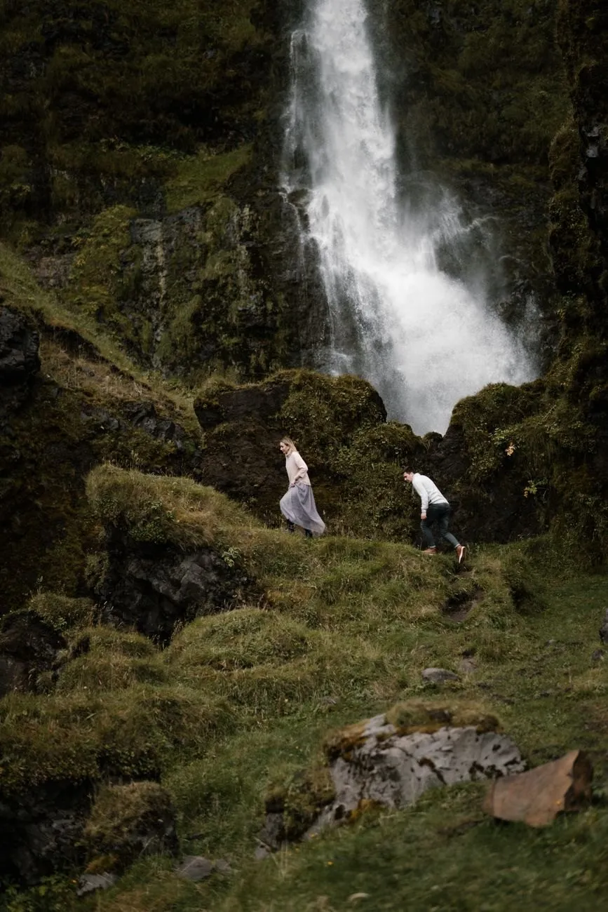 Two people climbing a mossy slope towards a waterfall