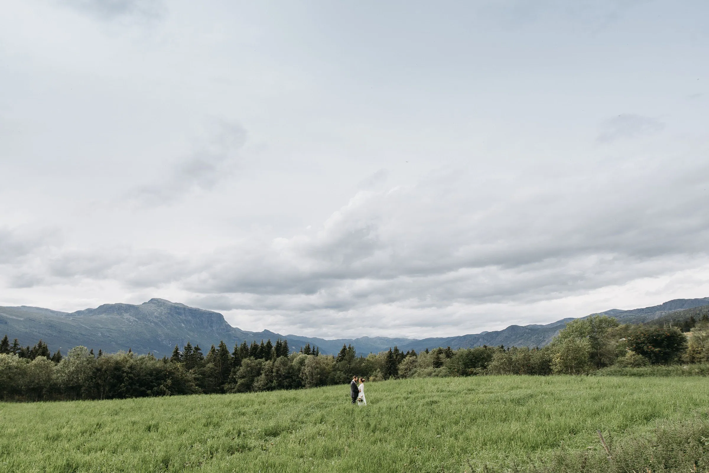 A couple standing together, tiny in a wide green hay meadow below a long mountain ridge in Hemsedal, Norway