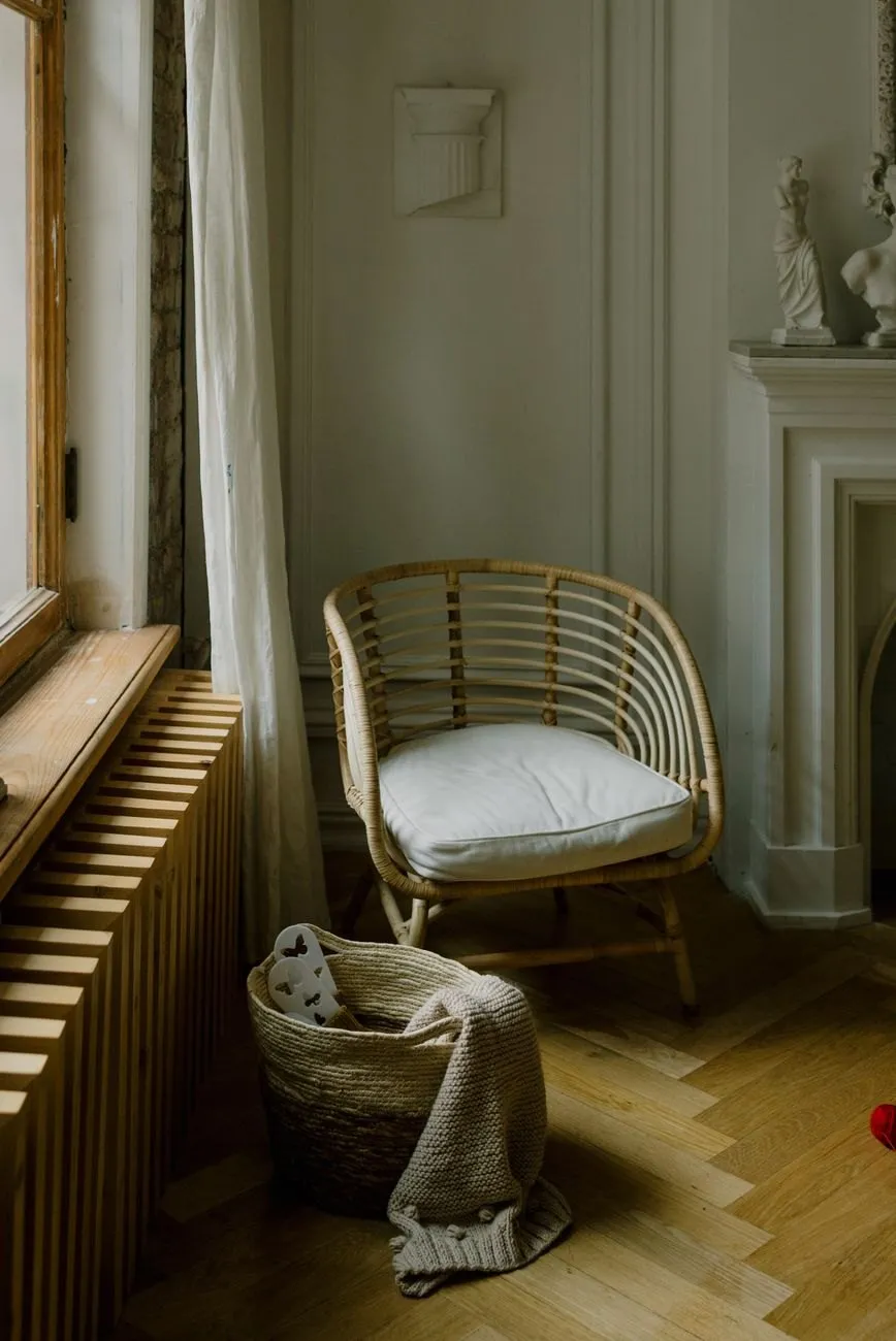 A rattan chair beside a tall window and a white fireplace with plaster casts on the mantel