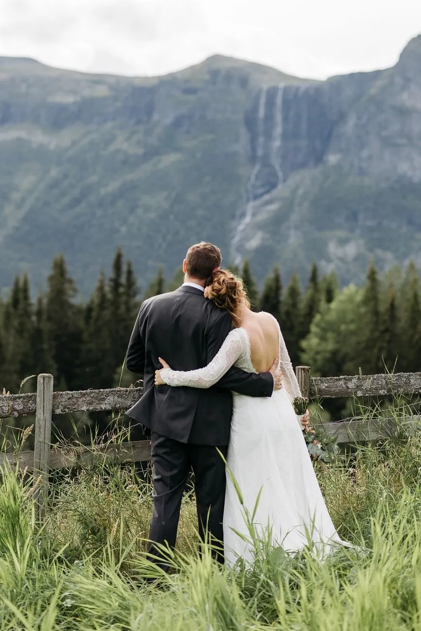 Ellie and Sam seen from behind, arm in arm at a wooden fence with a mountain wall and waterfall ahead