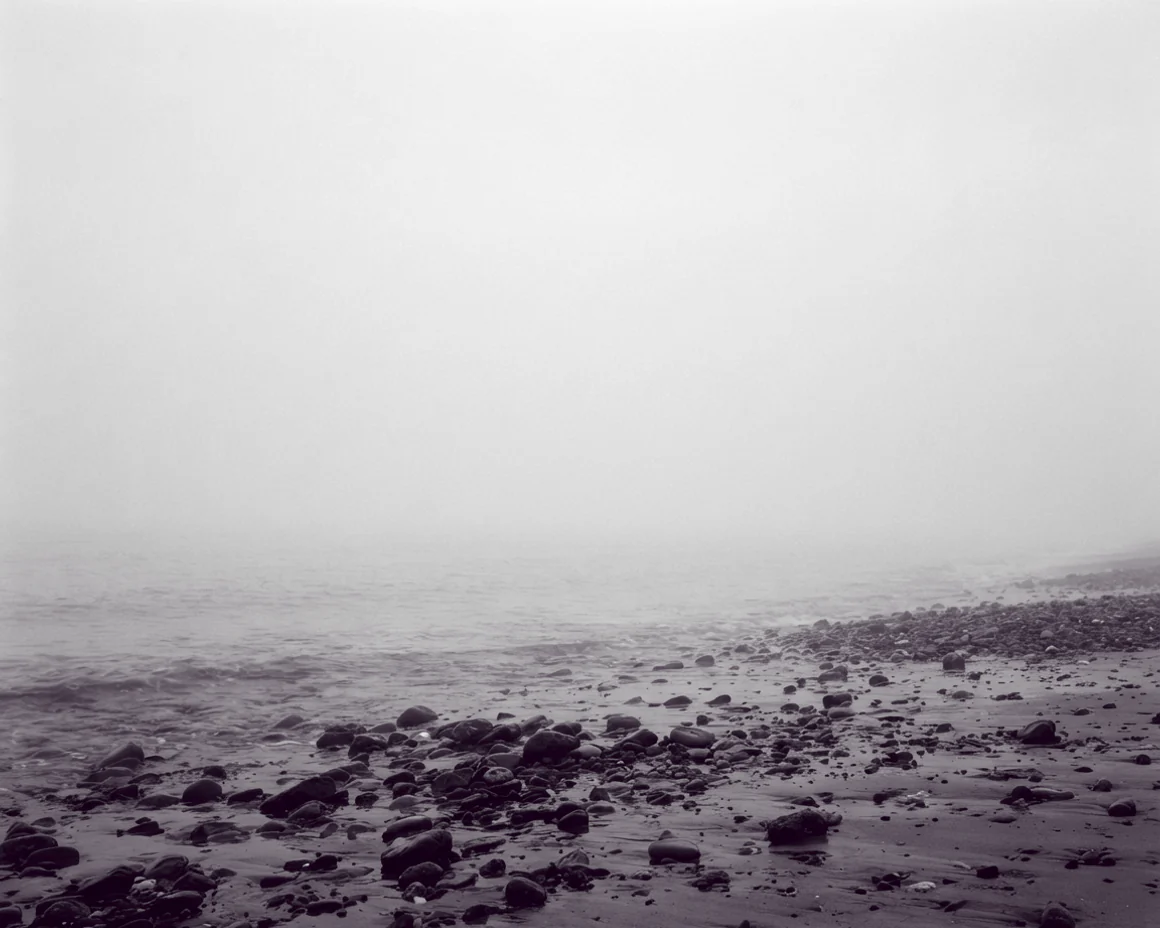 Dense fog reducing a shingle shoreline to a few dark stones in the foreground and pale grey beyond.