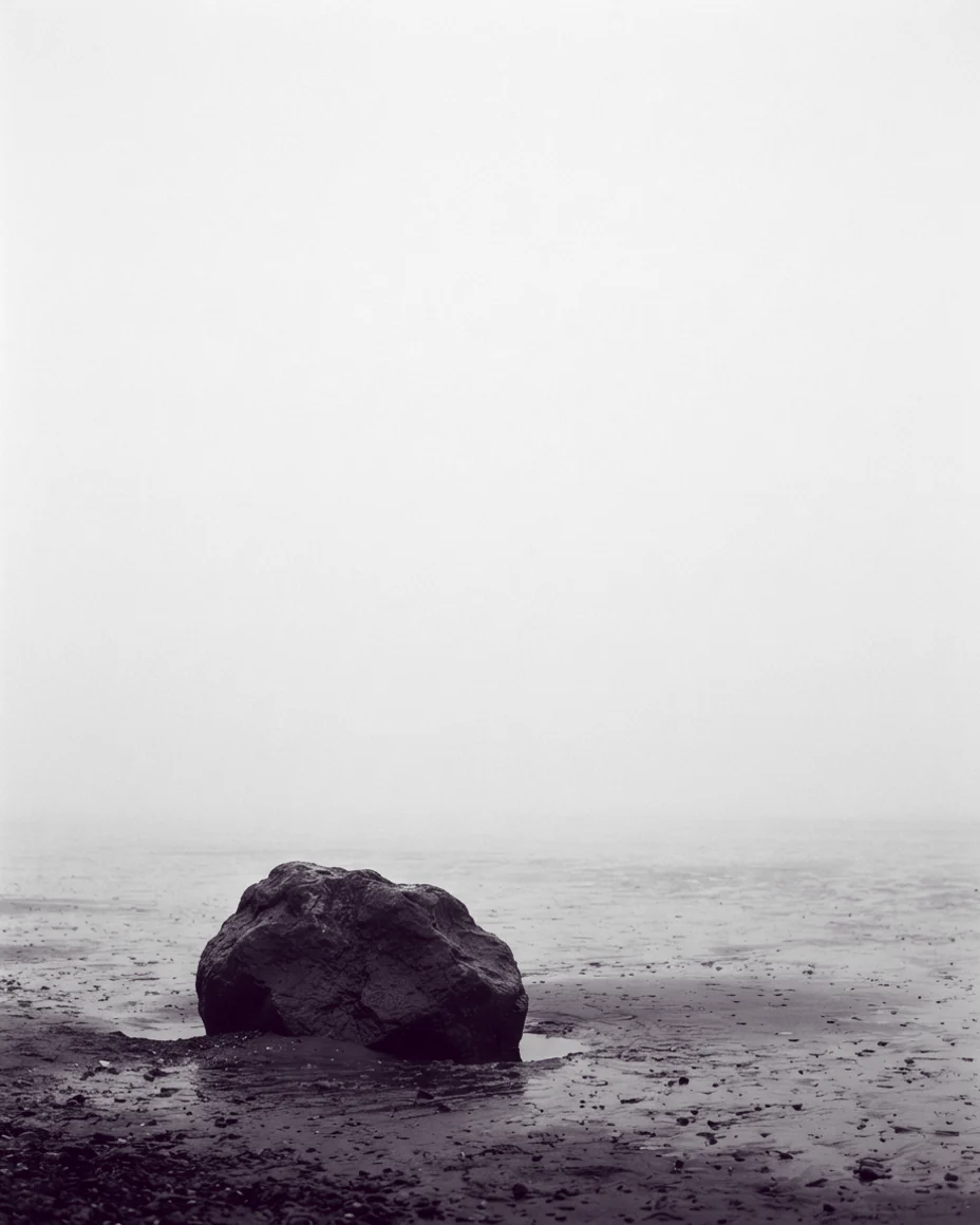 An upright high-key frame, one dark wet boulder low in the picture and empty pale fog above it.