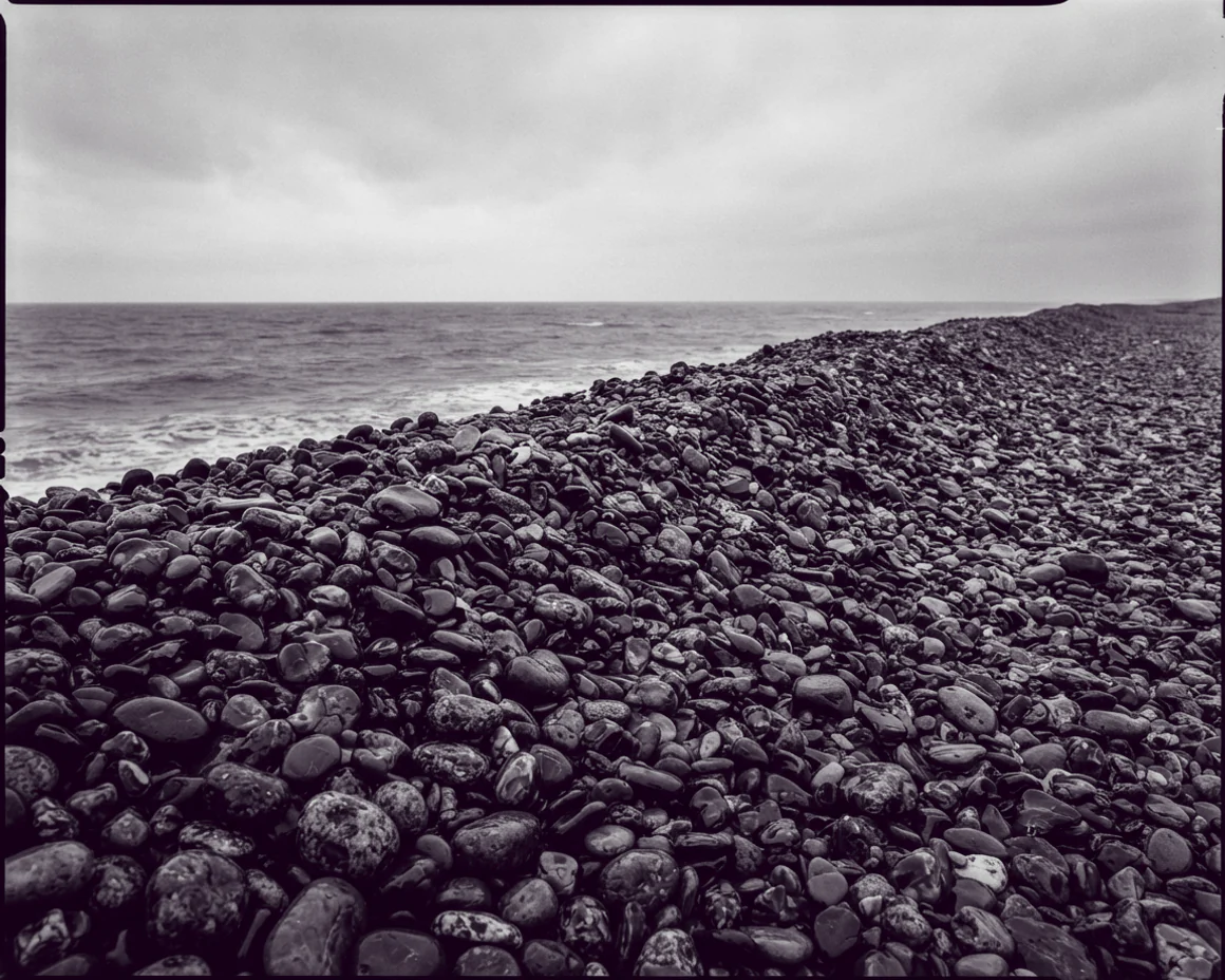 A storm-thrown shingle bank of wet rounded flint pebbles rising to a crest, flat grey sea beyond.